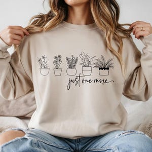 May include: Beige sweatshirt with a black line drawing of five potted plants. The text "just one more" is written in a cursive font below the plants. The sweatshirt has long sleeves and a crew neck.
