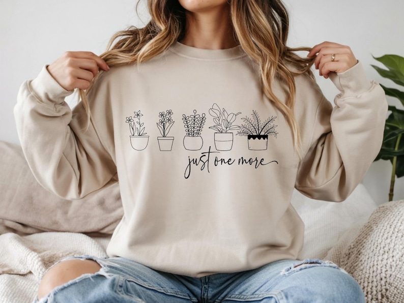 May include: Beige sweatshirt with a black line drawing of five potted plants. The text "just one more" is written in a cursive font below the plants. The sweatshirt has long sleeves and a crew neck.