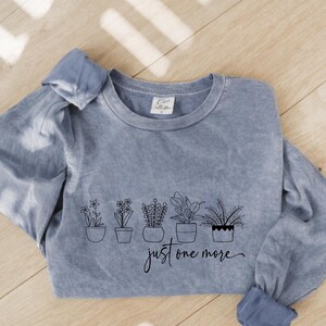 May include: A blue long-sleeve shirt with a crew neck. The shirt features a black line drawing of potted plants and the phrase "just one more" in a script font. The shirt has a faded, vintage look.