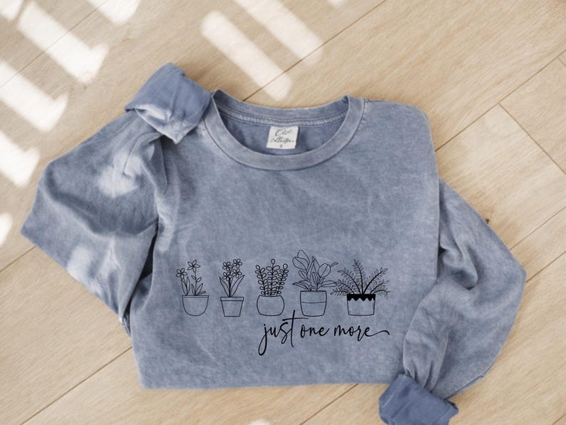May include: A blue long-sleeve shirt with a crew neck. The shirt features a black line drawing of potted plants and the phrase "just one more" in a script font. The shirt has a faded, vintage look.