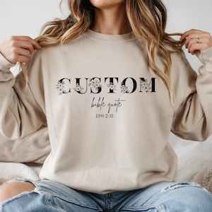 May include: Beige sweatshirt with the words "CUSTOM bible quote" in black, with floral accents. The text "EPH 2:10" is below the quote. The sweatshirt is a neutral colour and has long sleeves.