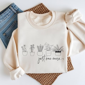 May include: Cream-colored sweatshirt with a crew neck and long sleeves. The front features a black line drawing of five potted plants with the text "just one more" in a script font. Blue jeans are visible on the left.