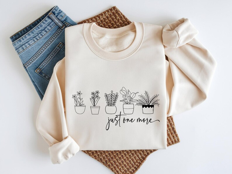 May include: Cream-colored sweatshirt with a crew neck and long sleeves. The front features a black line drawing of five potted plants with the text "just one more" in a script font. Blue jeans are visible on the left.