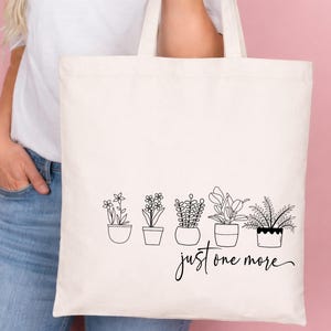 May include: Cream-colored tote bag featuring a black line drawing of five potted plants. The phrase "just one more" is written in a cursive font below the plants. The bag has two handles and is held against a pink background.
