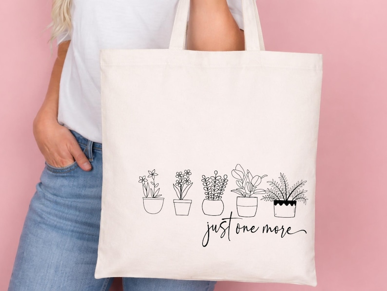May include: Cream-colored tote bag featuring a black line drawing of five potted plants. The phrase "just one more" is written in a cursive font below the plants. The bag has two handles and is held against a pink background.