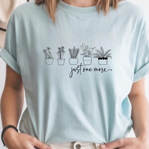 May include: A light blue t-shirt featuring a line drawing of five potted plants. The text "just one more" is written in a cursive font below the plants. The shirt has rolled-up sleeves.