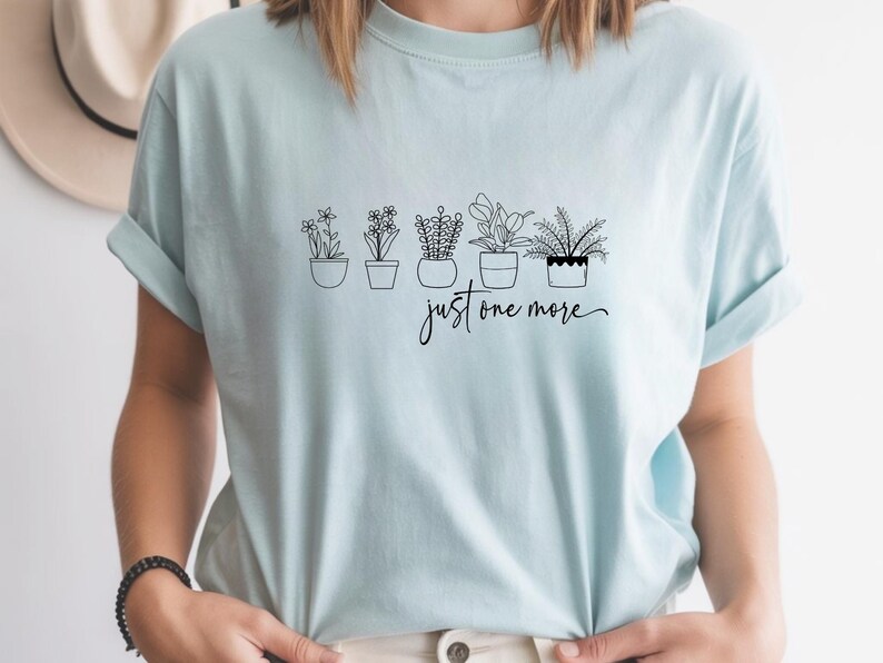 May include: A light blue t-shirt featuring a line drawing of five potted plants. The text "just one more" is written in a cursive font below the plants. The shirt has rolled-up sleeves.