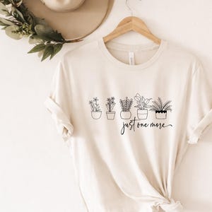 May include: Cream-colored t-shirt featuring a line drawing of five potted plants with the handwritten text "just one more" in black. The shirt is displayed on a wooden hanger.