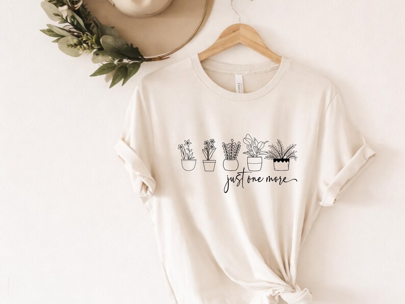 May include: Cream-colored t-shirt featuring a line drawing of five potted plants with the handwritten text "just one more" in black. The shirt is displayed on a wooden hanger.