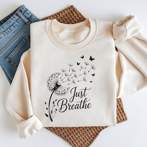 Op de afbeelding: Crèmekleurige sweatshirt met een zwart paardenbloemontwerp en de tekst "Just Breathe". De paardenbloem waait weg met vlinders. Een blauwe spijkerbroek en een bruine geweven mat zijn op de achtergrond.