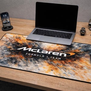May include: A desk setup featuring a laptop, phone, and accessories. A large mousepad with a watercolor design and the McLaren Formula 1 Team logo dominates the scene. A black mouse, watch, and skull are also visible.