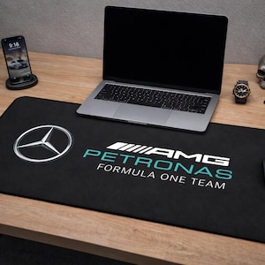 May include: Black desk mat with the Mercedes-Benz logo and "AMG Petronas Formula One Team" text in white and teal. A laptop, phone, watch, and other items are on the wooden desk. The mat measures approximately 80 cm in length.