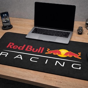 Peut inclure: Tapis de souris rectangulaire noir avec le logo Red Bull Racing en rouge et blanc. Un ordinateur portable, un téléphone, une montre et d'autres objets sont sur un bureau en bois.