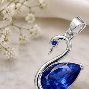 May include: A silver-toned metal pendant shaped like a swan, featuring a large, pear-shaped deep blue gemstone as the body and a small, round blue stone set as the eye. The pendant includes a polished metal bail at the top for chain attachment.