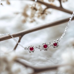 Könnte beinhalten: Eine zarte Silberkette mit drei Blütenanhängern, jeweils mit einem leuchtend roten Edelstein in der Mitte. Das florale Design ist durch eine filigrane Kette verbunden und schafft ein elegantes und feminines Schmuckstück.