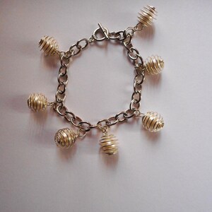 May include: Silver-toned bracelet featuring a chain design with multiple spherical cage-like charms. The bracelet has a toggle clasp. The charms are made of thin, coiled metal, creating a unique, openwork design. The bracelet is displayed on a white surface.