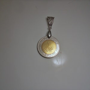 Op de afbeelding: Een zilveren hanger met een gouden munt met Arabische tekst en de woorden "ONE POUND 1411 H" rond de rand.