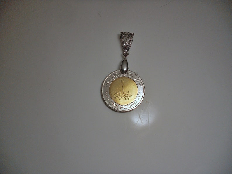 Op de afbeelding: Een zilveren hanger met een gouden munt met Arabische tekst en de woorden "ONE POUND 1411 H" rond de rand.