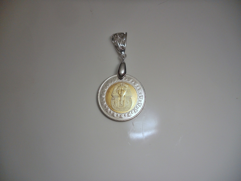 Op de afbeelding: Een zilveren hanger met een rond gouden sieraad met een buste van een Egyptische farao. De hanger is omringd door hi&euml;rogliefen.