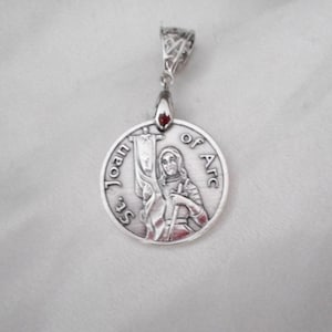 May include: A circular silver-toned metal pendant featuring a relief image of a figure holding a banner and a sword. The text St. Joan of Arc is embossed along the curved edge. The pendant includes a decorative filigree bail at the top.