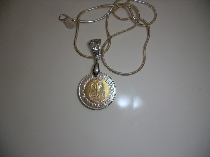Op de afbeelding: Een zilveren ketting met een ronde hanger met een gouden Egyptisch faraosymbool.
