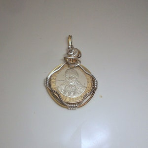 May include: A silver and gold coin pendant with a portrait of a man on the front. The coin is surrounded by a silver wire frame and has the text "REPUBLICA DE CHILE" and "LOS ORIGINARIOS" on it.