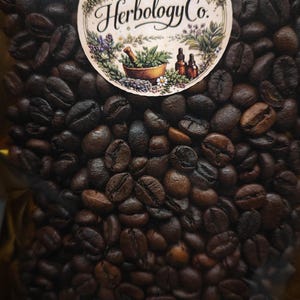 Könnte beinhalten: Nahaufnahme eines transparenten Beutels gefüllt mit gerösteten Kaffeebohnen. Ein rundes Etikett mit dem Text "Herbology Co." ist sichtbar, umgeben von floralen Illustrationen und einem Mörser und Stößel. Die Bohnen sind dunkelbraun.