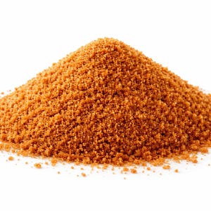 Op de afbeelding: Een berg bruine kristalsuiker tegen een witte achtergrond. De suiker heeft een grove textuur en een warme, roodbruine kleur. De afbeelding is een close-up, die de korrelige details benadrukt.