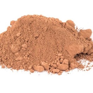 May include: A mound of brown powder with a fine texture and small, irregular clumps. The powder is a warm, earthy tone, set against a plain white background.