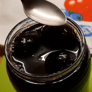 May include: A clear glass jar filled with dark, viscous liquid and several whole dark fruits submerged within. A silver-coloured metal spoon is positioned directly above the open top of the jar, reflecting light on its smooth, curved surface.