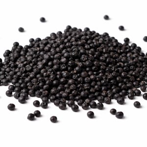May include: A mound of black peppercorns against a white backdrop. The peppercorns are small, spherical, and dark, with a slightly textured surface. Some peppercorns are scattered around the main pile.
