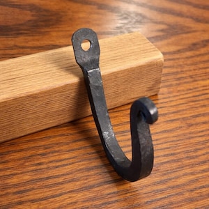 Puede incluir: Un gancho de metal negro forjado a mano con un diseño curvo. El gancho tiene un lazo en la parte superior para colgar y un extremo rizado. Se muestra sobre un fondo de madera, lo que sugiere un estilo rústico.