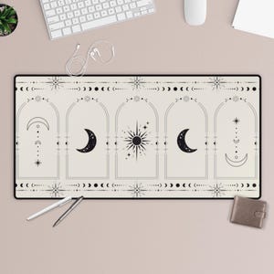 Puede incluir: Una alfombrilla de escritorio rectangular con un fondo crema y un diseño celestial negro. El diseño presenta lunas, estrellas y soles dentro de marcos arqueados. Un bolígrafo blanco, un bolígrafo plateado y una pequeña cartera marrón están sobre la alfombrilla.