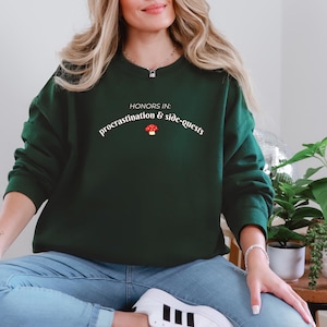 May include: Dark green sweatshirt with the text "HONORS IN: procrastination & side-quests" in white, with a red mushroom graphic. The sweatshirt is a crew neck style.