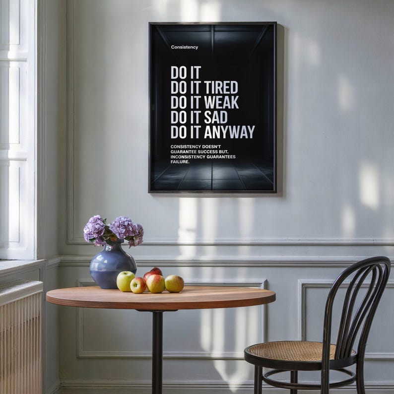 May include: A framed black and white motivational poster with the text "DO IT" and other phrases. Below the poster is a round wooden table with a blue vase of purple flowers and several apples. A black chair sits to the right.