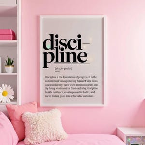 May include: A framed print with the word "discipline" in bold black letters. The definition of discipline is below the word. The print is in a white frame and hangs on a pink wall. The room has pink pillows and a white shelf.