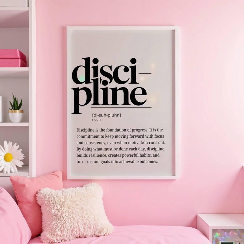 May include: A framed print with the word "discipline" in bold black letters. The definition of discipline is below the word. The print is in a white frame and hangs on a pink wall. The room has pink pillows and a white shelf.