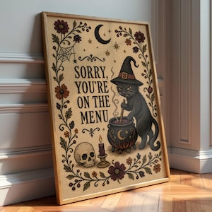 May include: A rectangular framed print on a tan background featuring a black cat wearing a witch hat, stirring a cauldron. The text reads, SORRY, YOU'RE ON THE MENU. The design includes a skull, a candle, a crescent moon, and floral borders.