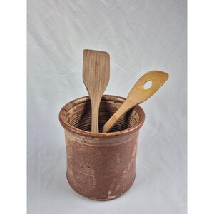 May include: A cylindrical brown ceramic crock with a speckled finish and ribbed interior texture. Inside the crock are two light-colored wooden kitchen utensils, one flat-edged spatula and one spoon with a circular hole in the center.