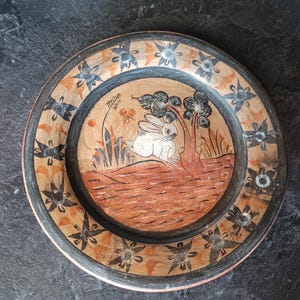 Puede incluir: Plato redondo de cerámica pintado a mano con una escena de conejo. El plato presenta una imagen central de un conejo blanco bajo un árbol, rodeado de patrones florales y geométricos en negro, naranja y marrón. El plato tiene un diámetro de unos 25 cm.