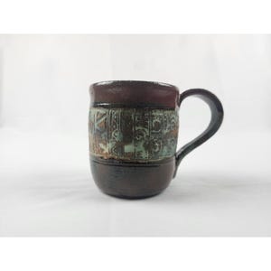 May include: A cylindrical ceramic mug with a dark brown glazed finish. The center of the mug features a wide, textured band with a light green, weathered patina and intricate, carved geometric patterns encircling the entire body.