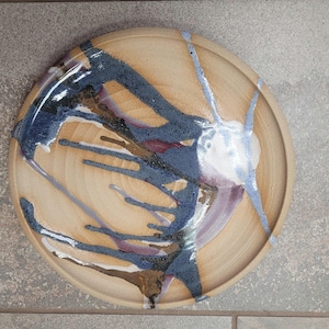 May include: A handmade ceramic plate with a beige base and abstract blue, brown, and white glaze design. The plate has a circular shape with a slightly raised rim, suitable for serving food or as a decorative piece.