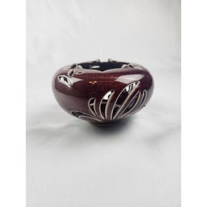 May include: A deep burgundy ceramic bowl featuring a glossy finish and intricate cut-out patterns along the sides. The bowl has a rounded, bulbous shape with a wide opening at the top, showcasing a dark interior and detailed carved leaf-like motifs.