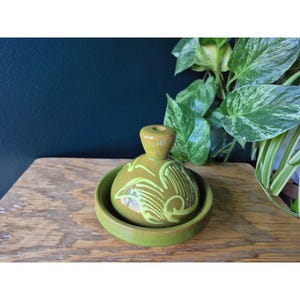 May include: A small, round, olive-green ceramic container with a matching lid. The lid features a decorative swirl design in a lighter green. The container sits on a wooden surface, with leafy green plants in the background.