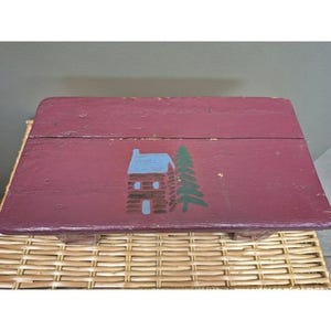 May include: A rectangular wooden step stool painted in a deep maroon color. The top surface features a hand-painted graphic of a small cabin with a light blue roof and a tall green pine tree standing directly beside the structure.