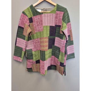 May include: A patchwork-style long-sleeve shirt in shades of green, pink, and purple. The shirt features a variety of patterned squares, creating a unique and textured design. The shirt is hanging on a wooden hanger.