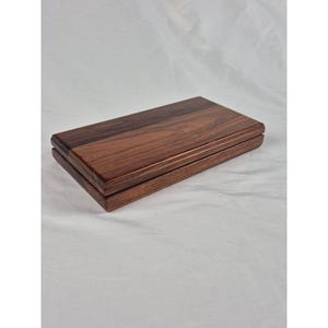 May include: A rectangular wooden box with a smooth, polished finish. The lid features a rich, dark brown wood grain pattern with visible horizontal streaks and natural variations in color, showcasing a deep mahogany and chestnut tone throughout.