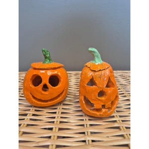 May include: Two orange ceramic jack-o'-lanterns with green stems. The left pumpkin is round with a wide, smiling face and circular eyes. The right pumpkin is taller and narrower with a jagged, triangular-eyed face and a wide, toothy grin.