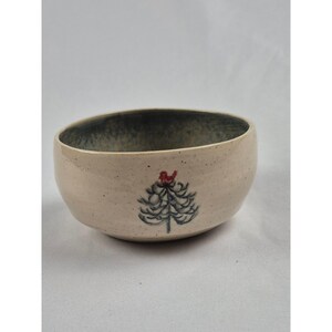 May include: A speckled beige ceramic bowl with a rounded shape and a dark interior. The exterior features a hand-painted dark gray evergreen tree illustration topped with a small, vibrant red bird perched at the very peak of the tree design.