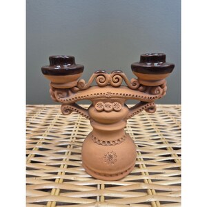 May include: A terracotta double candle holder featuring a central pedestal base with intricate stamped circular patterns and scrollwork details. The two candle cups are finished with a smooth, dark brown glazed rim, contrasting with the matte clay.